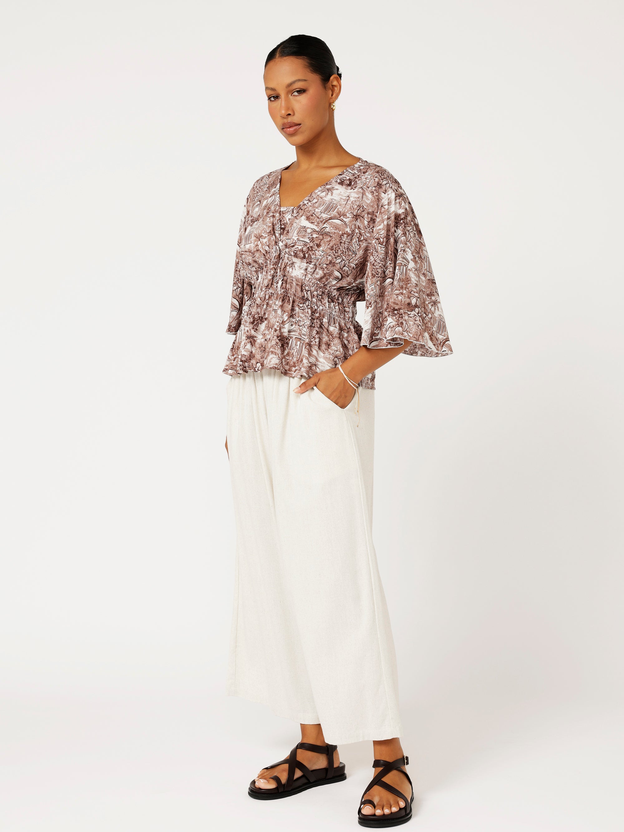 side view of brown Toile de Jouy top with fluted sleeves and ruched waist styled with natural linen pants